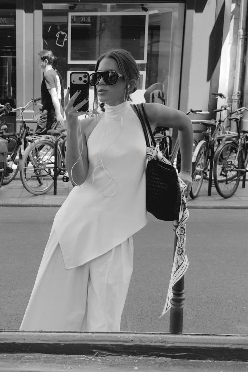 Belen Gonzalez Castellanos taking a mirror selfie in a white asymmetric top and wide-leg trousers, carrying a black woven bag with a printed scarf
